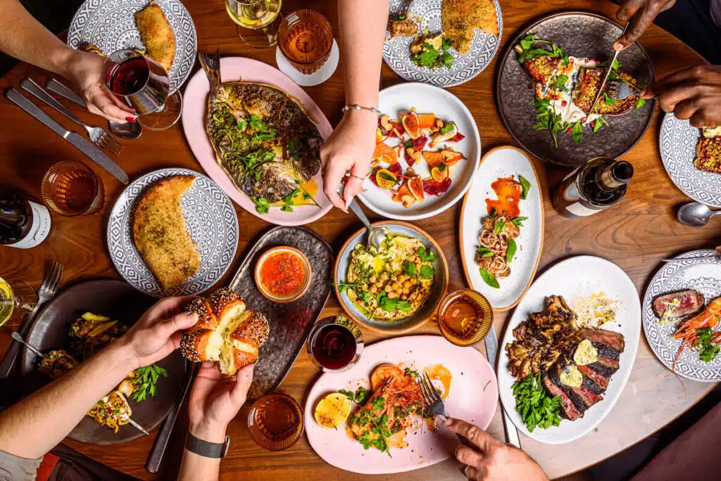 People sharing a variety of colorful dishes and drinks on a wooden table, seen from above.