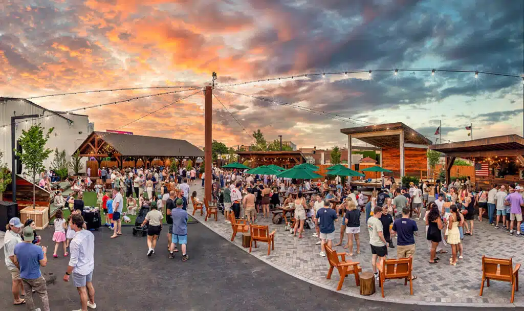 2026 Boston Travel Guide. Large crowd gathers outdoors at sunset, socializing around tables and under string lights in a lively setting.