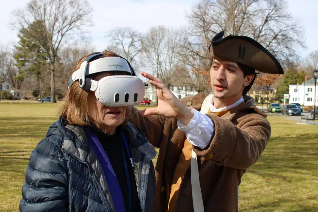 A man in colonial attire helps a woman wear a virtual reality headset outdoors in a park.