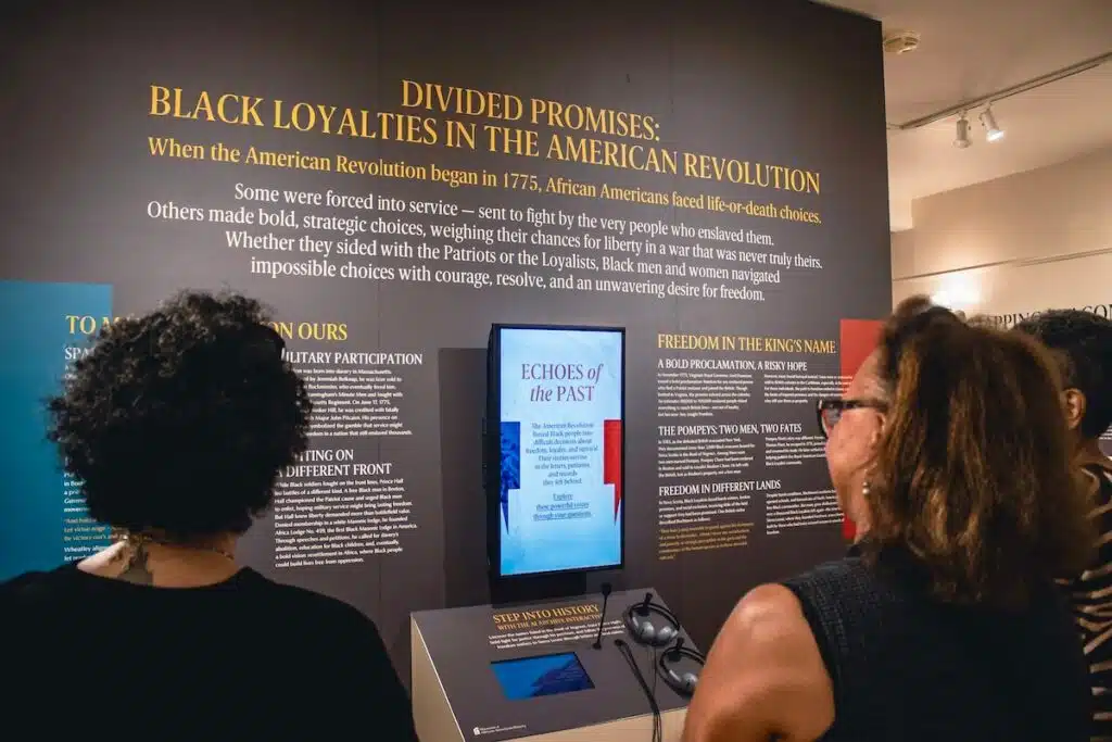 Three people view a museum exhibit about Black Loyalists in the American Revolution titled "Divided Promises.