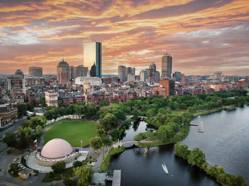 A city skyline at sunset with a river, park, and dome-shaped building in the foreground.
