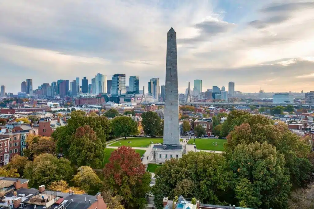 A tall stone obelisk stands in a park with a city skyline in the background under a cloudy sky.