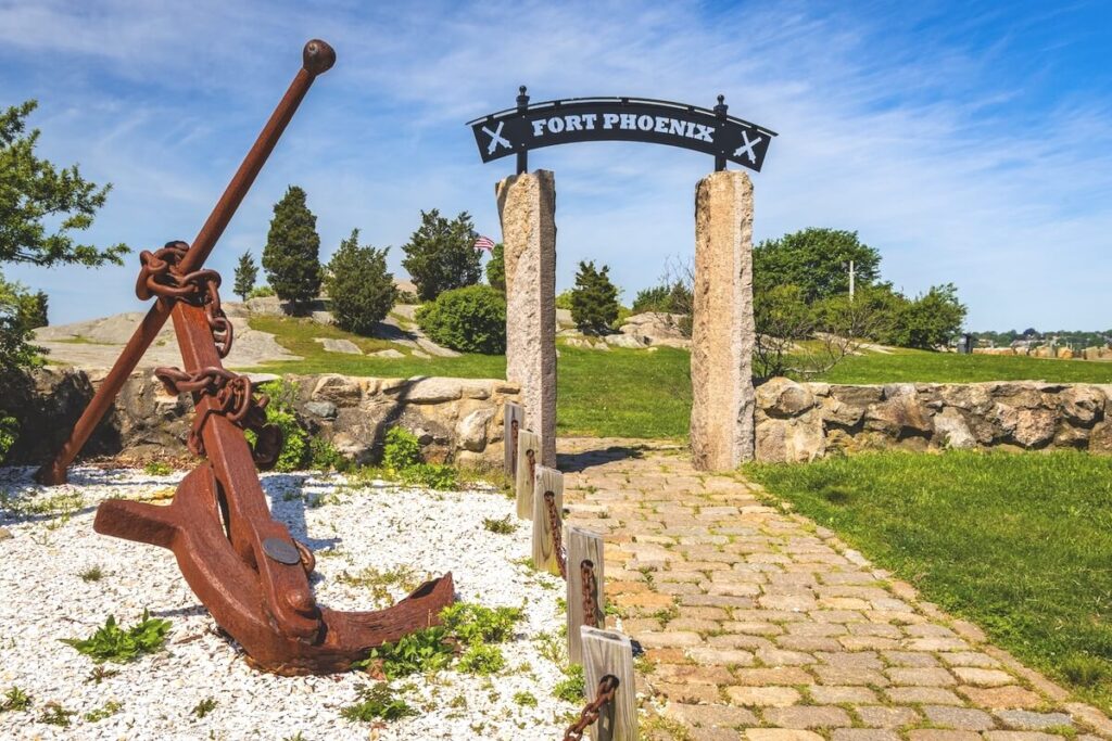 Large anchor and stone path leading to "Fort Phoenix" sign under a blue sky, surrounded by grass and trees.