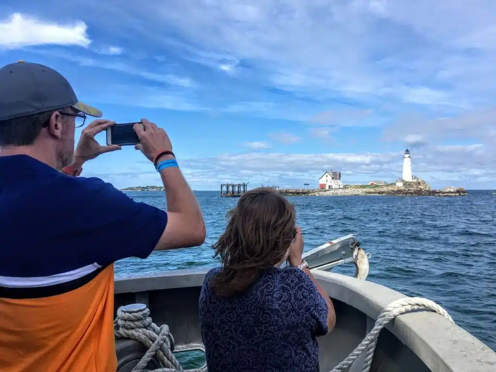 Two people on a boat take photos of a distant lighthouse on a small island under a blue sky.