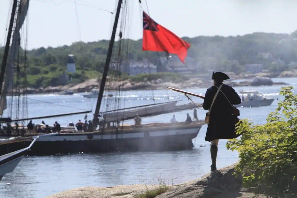 A person in colonial attire holds a musket by the water with a historical ship and red flag in the background.