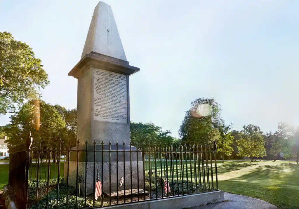 Tall stone monument with an inscription, surrounded by a black iron fence, in a sunny park with trees and flags.