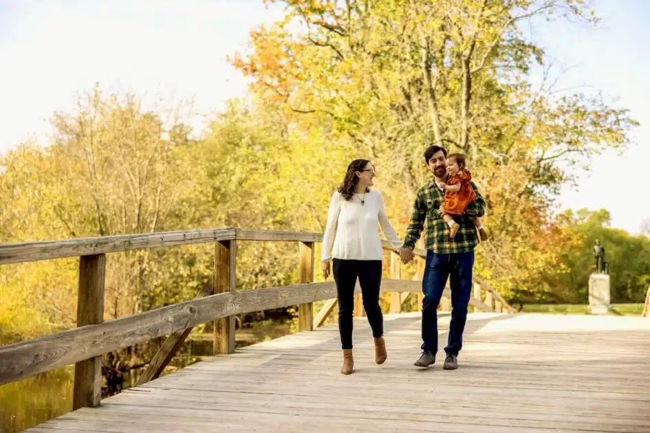 A couple walks hand in hand on a wooden bridge while a man holds a small child on a sunny autumn day.