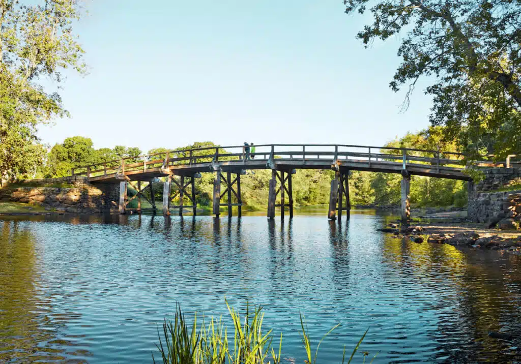 Wooden bridge over a calm river, surrounded by green trees and blue sky on a sunny day.