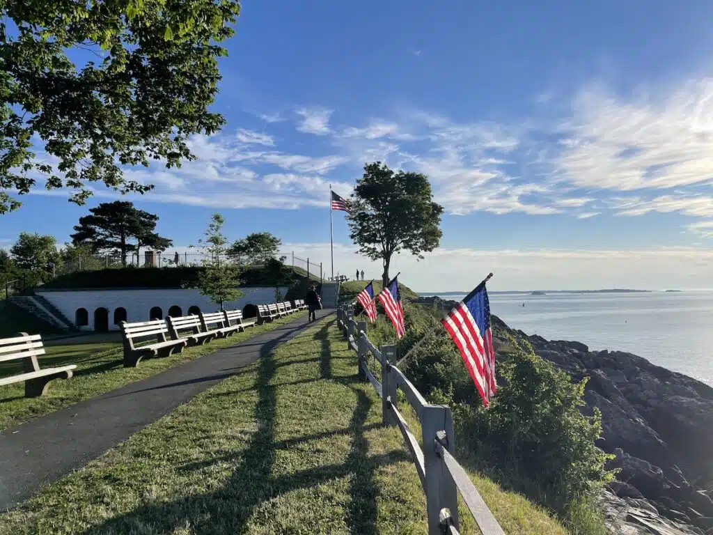 A coastal pathway with American flags, benches, trees, and ocean views under a clear blue sky.
