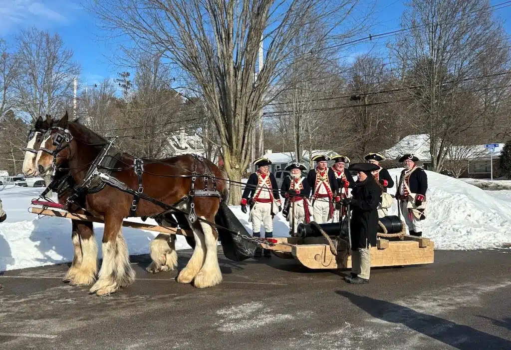 Two horses pull a wooden sled carrying a cannon; people in historical uniforms stand nearby in the snow.