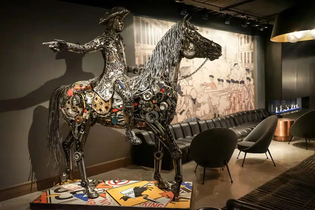Sculpture of a rider on a horse made from metal parts, displayed in a modern lounge with armchairs.