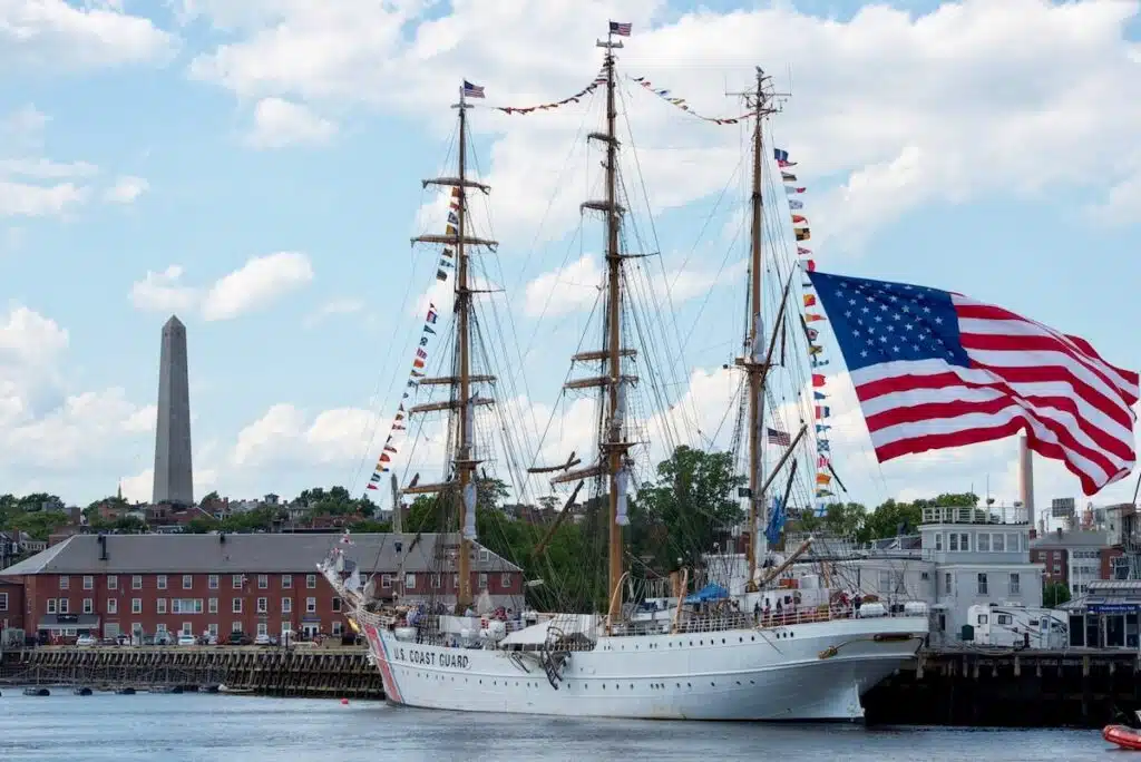 A tall ship with an American flag docked by historic buildings and a tall obelisk in the background.