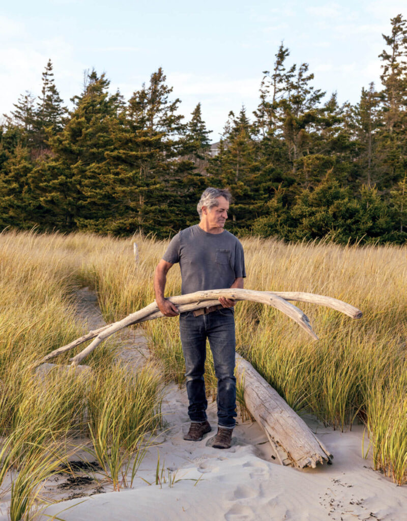 Man in a gray shirt carries driftwood on a sandy path with tall grass and pine trees in the background.