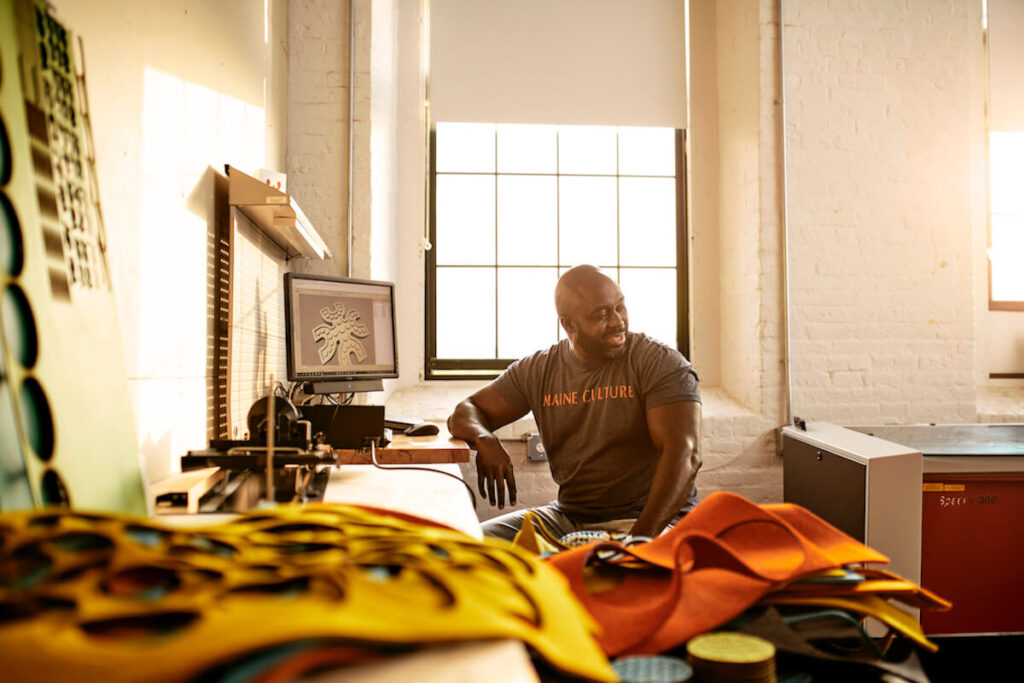 Man in a t-shirt sitting at a desk in a sunlit studio with colorful fabrics and a computer.