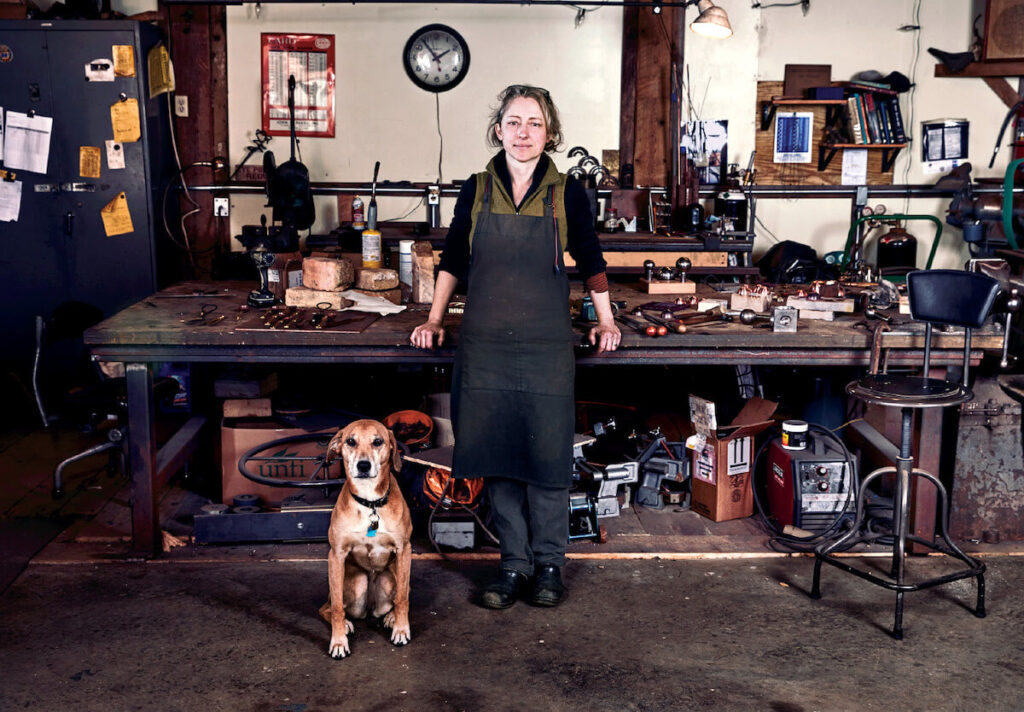 A person in a workshop stands behind a workbench with tools, accompanied by a brown dog sitting in front.