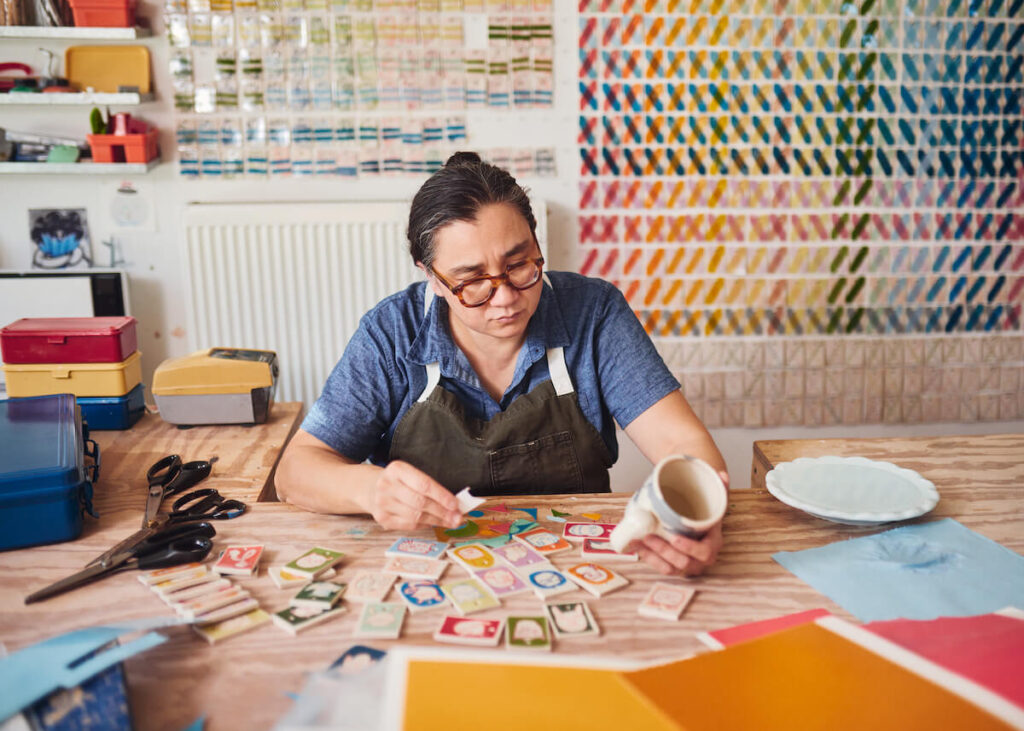 A person sits at a table arranging colorful patches, surrounded by craft supplies in a creative studio.