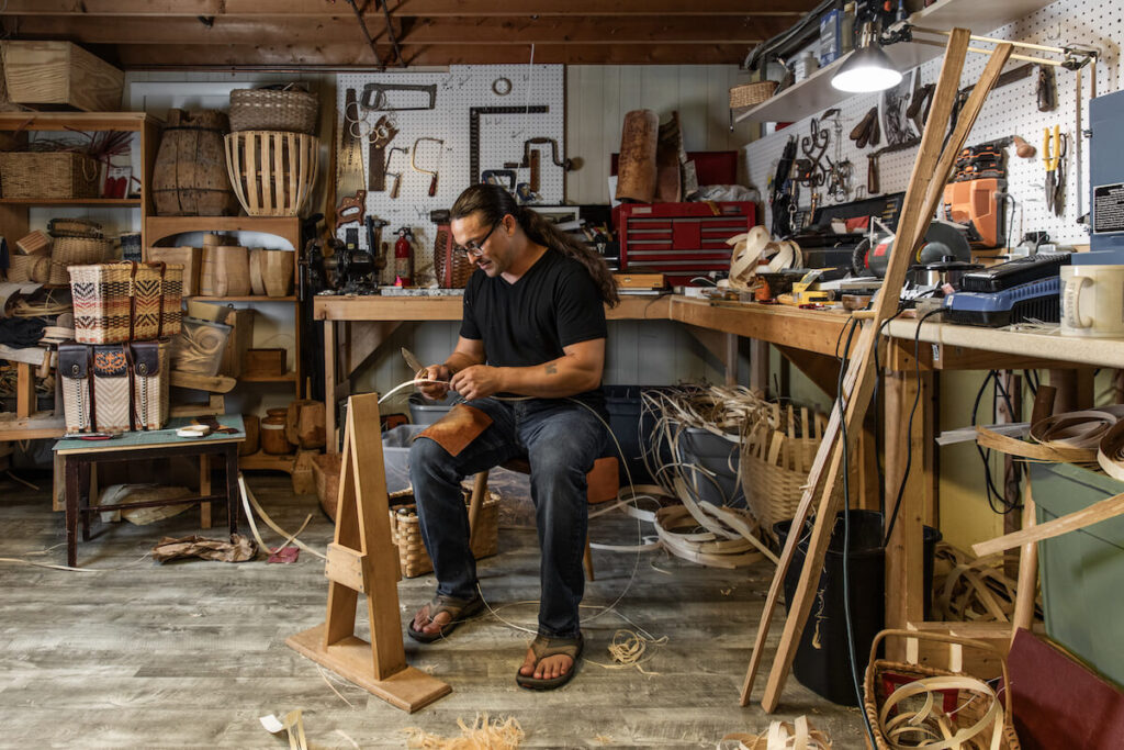 A man weaves a basket by hand in a workshop filled with tools, baskets, and weaving materials.