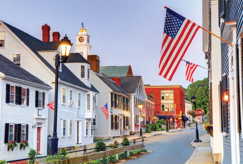 American flags hang from historic buildings lining a quiet street in a small New England town at dusk.
