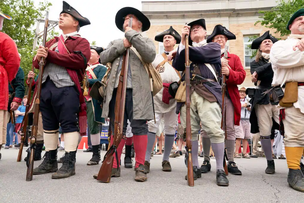 A group of people in colonial costumes hold muskets during a historical reenactment outdoors.