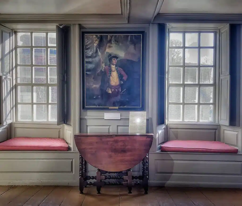 A portrait hangs between two window seats with red cushions and a folded wooden table below in a sunlit room.