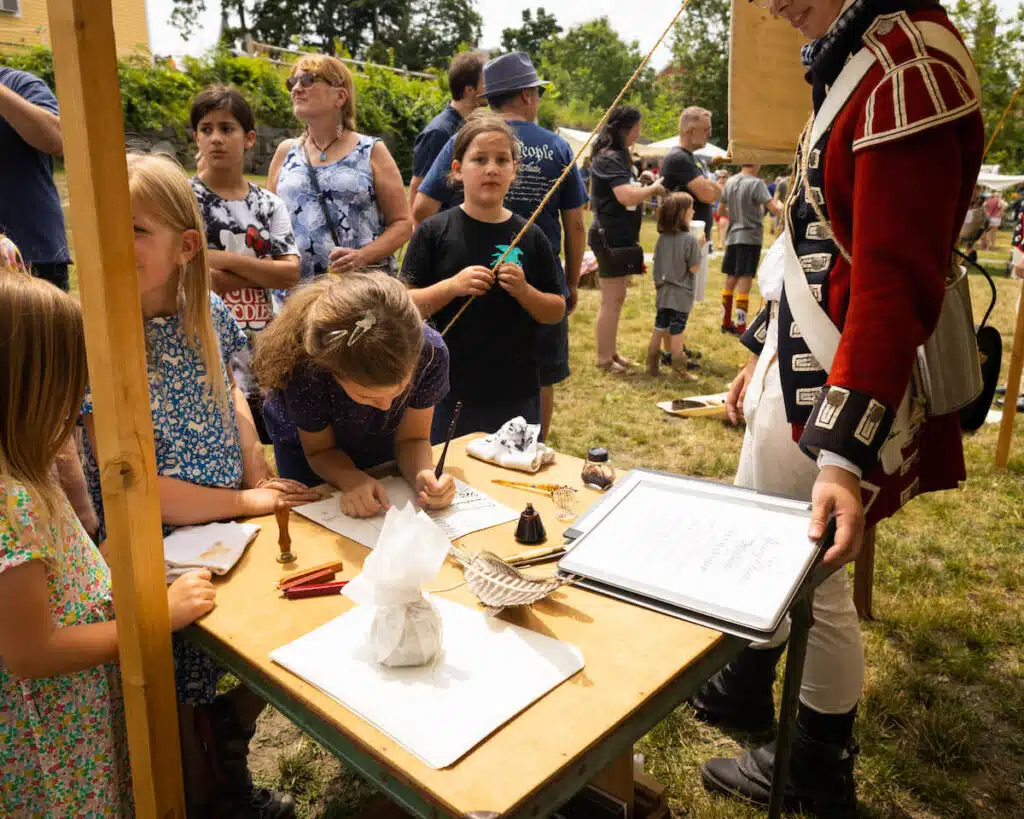 Children write at a wooden table outdoors during a historical reenactment with a reenactor in period costume.