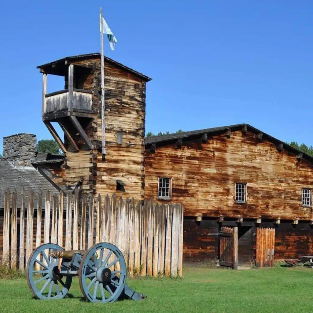 Old wooden fort with a watchtower, tall fence, waving flag, and a cannon on green grass under blue sky.