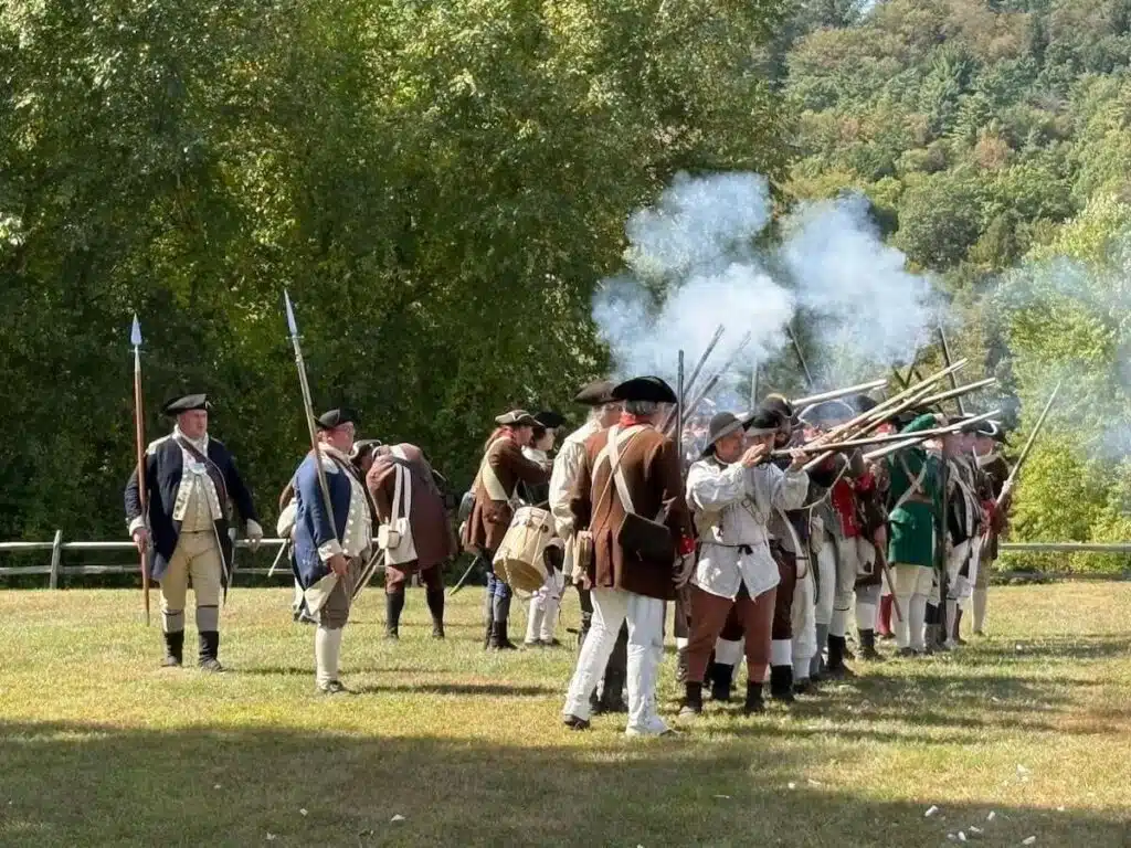 People in colonial-era clothing reenact a historical battle, firing muskets outdoors on a sunny day.