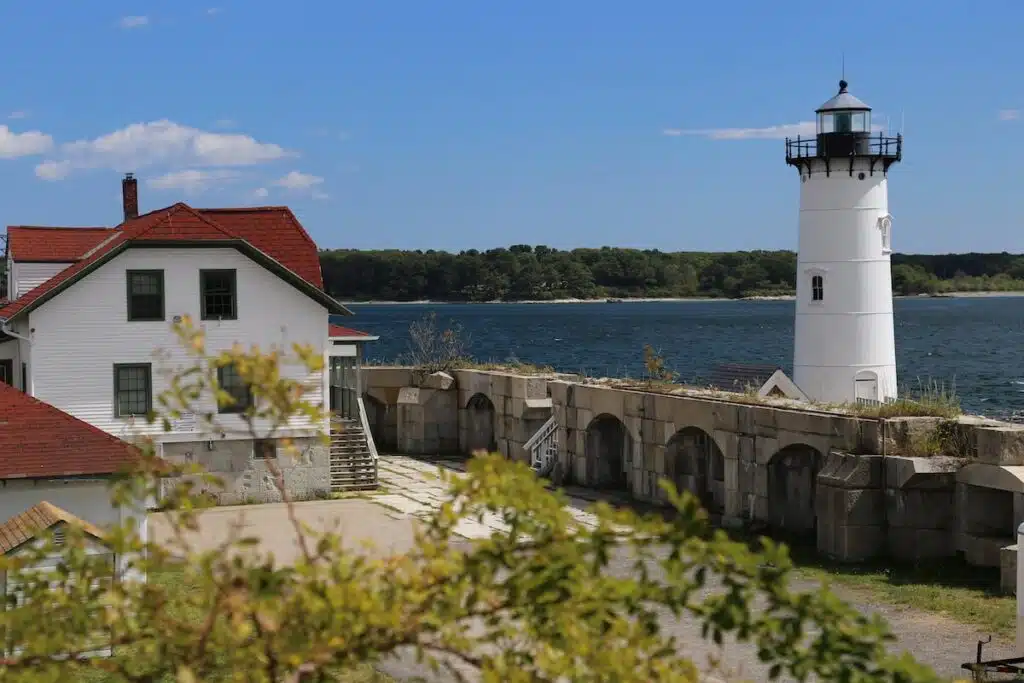 White lighthouse and house with red roof by the water, with trees and blue sky in the background.