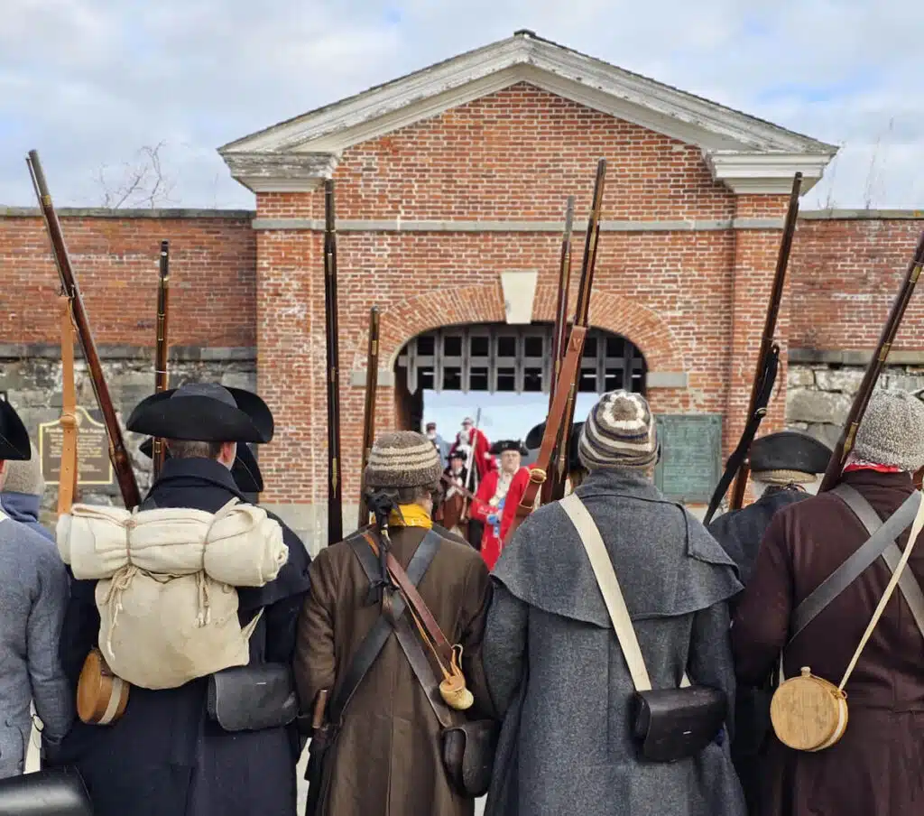 A group of reenactors in historical clothing face a brick fort entrance, holding muskets and gear.