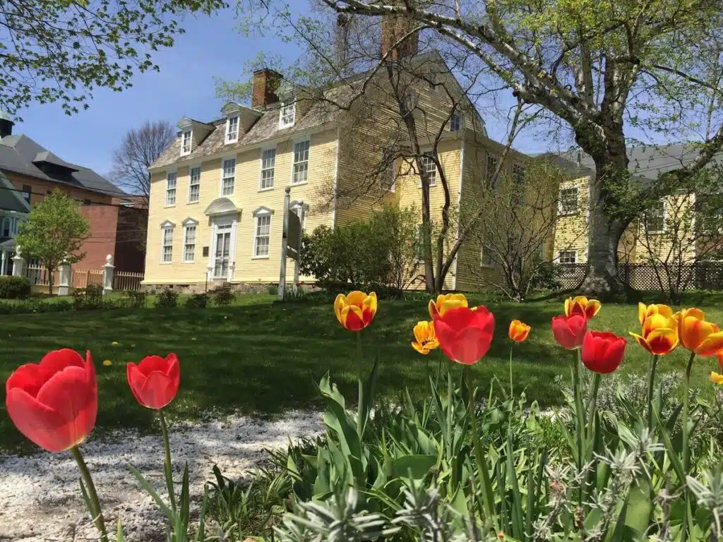Red and yellow tulips bloom in front of a large, pale yellow historic house on a sunny spring day.