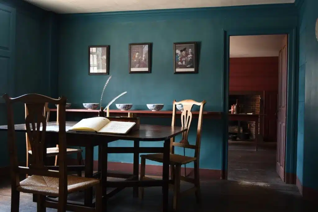 A vintage study room with blue walls, a table, chairs, an open book, and framed art on the wall.