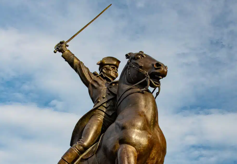 Bronze statue of a man on horseback holding a raised sword against a partly cloudy sky.
