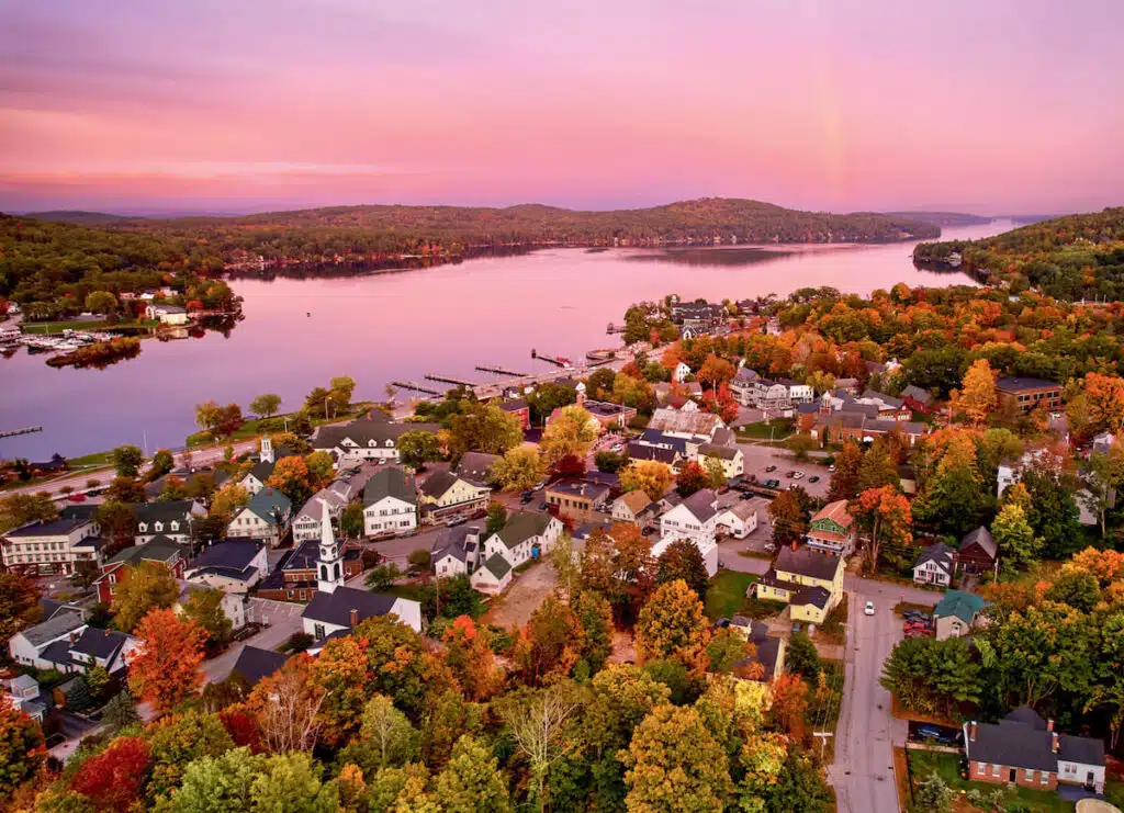 Aerial view of a small town by a lake with colorful autumn trees under a pink sunset sky.
