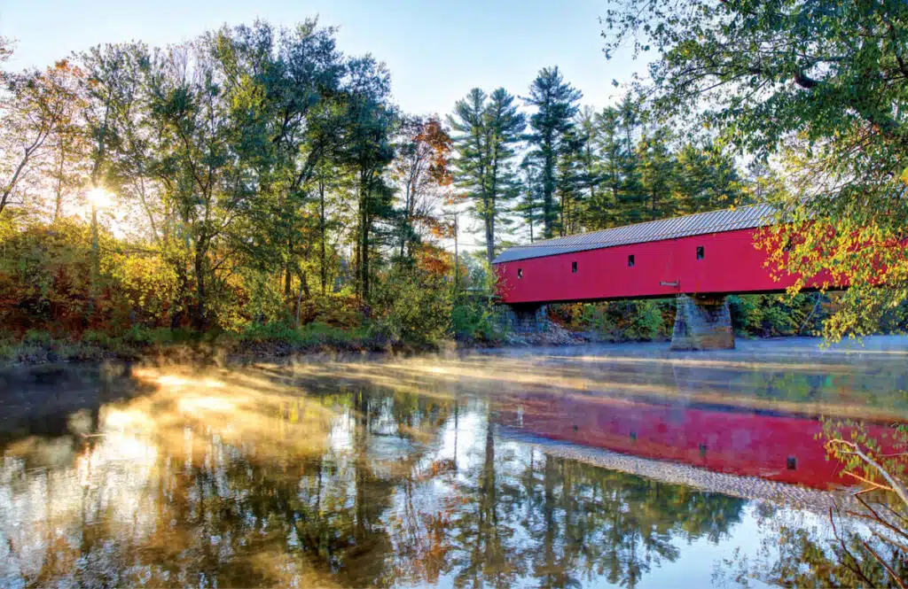 A red covered bridge spans a calm river surrounded by trees with sunlight streaming through.