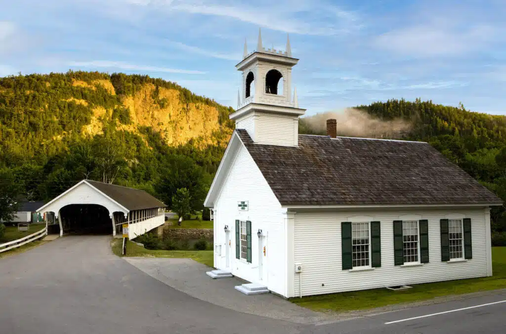 White church with green shutters near a covered bridge and forested hill in the background, under a clear sky.