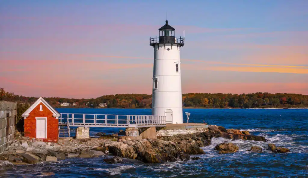 White lighthouse and small red building on rocky coast with blue water, trees, and a colorful sky at sunset.