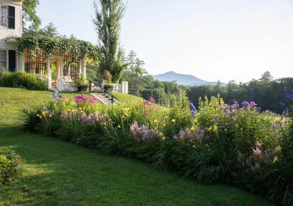 A lush garden with colorful flowers, green lawn, porch, and distant mountain under a clear sky.