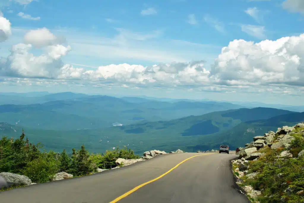 Car driving up a mountain road with scenic views of green hills, distant mountains, and a partly cloudy sky.