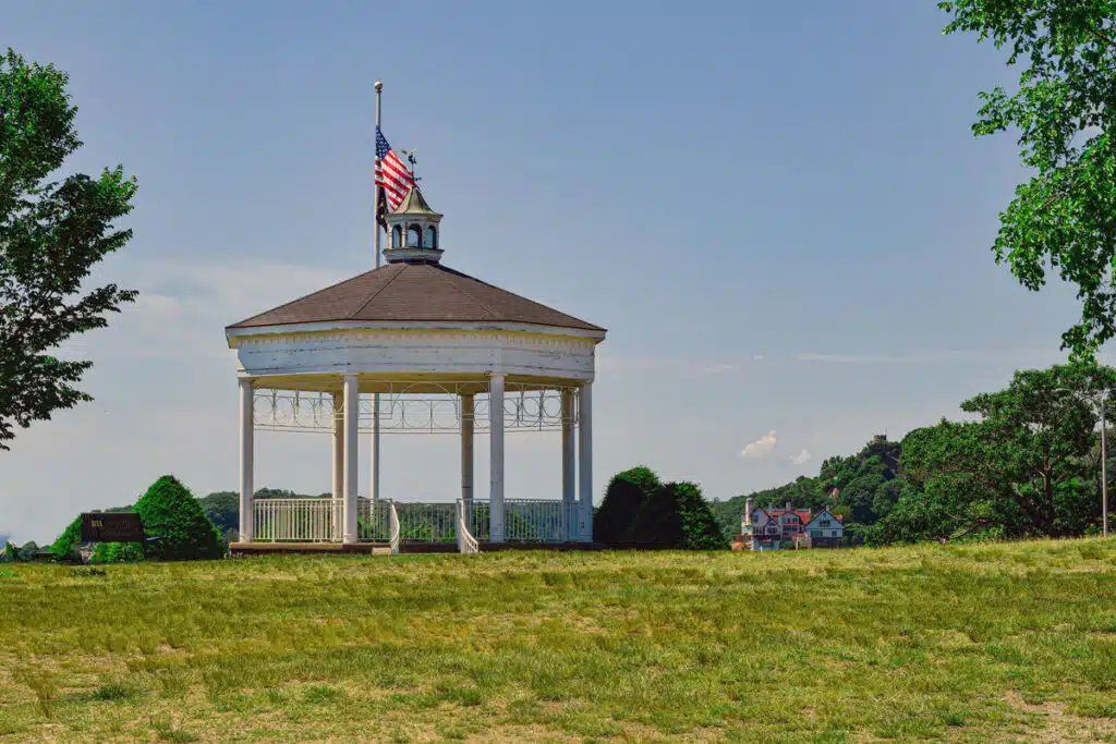 A white gazebo with an American flag stands on a grassy hill, with trees and houses in the background.