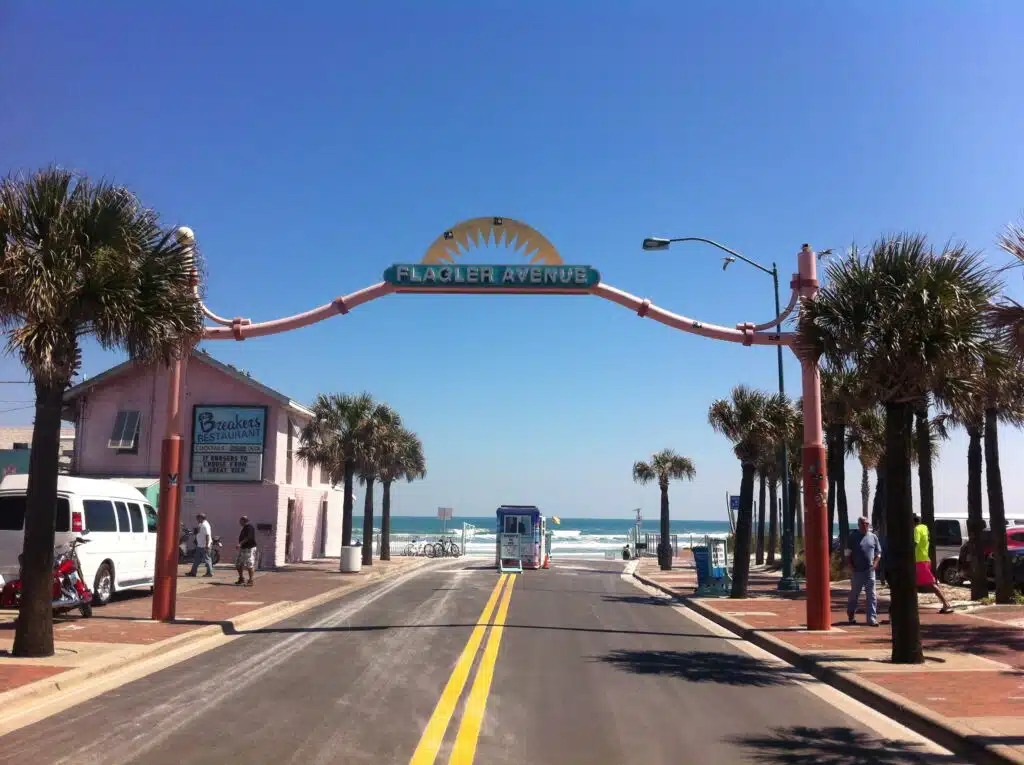 Palm-lined street leading to the beach with a "Flagler Avenue" archway and people walking near the ocean.