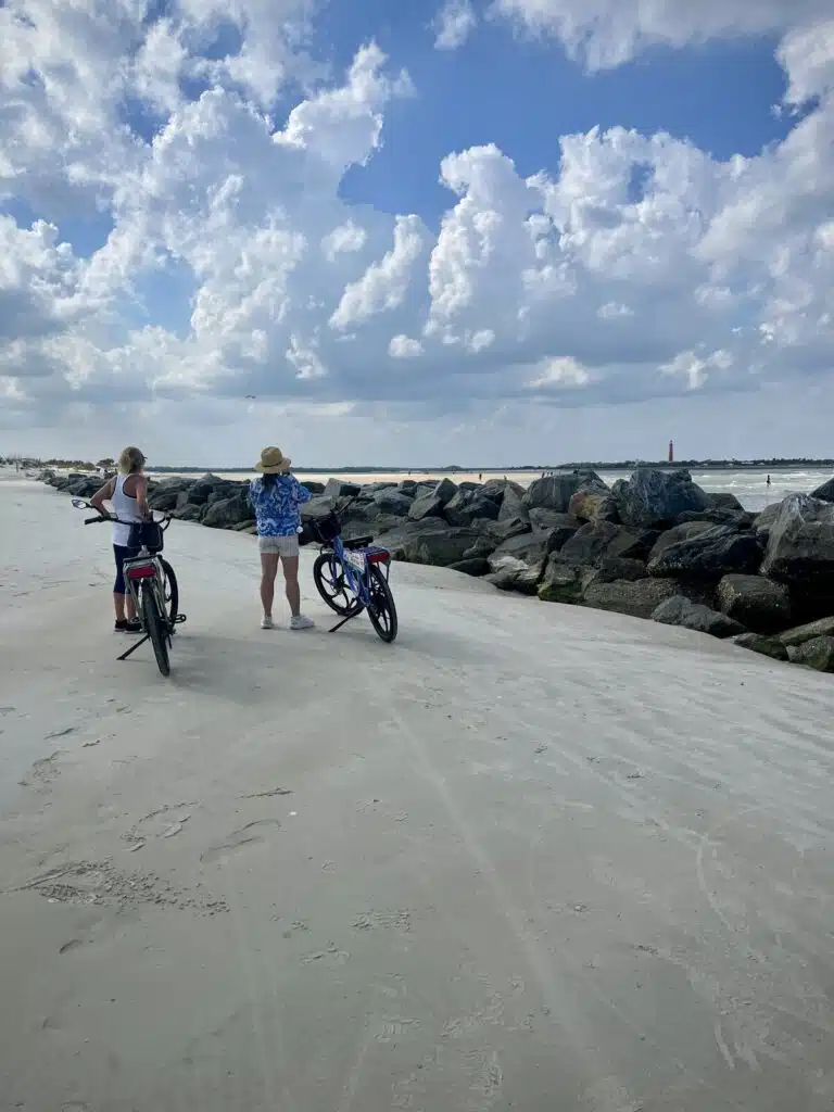 Two people with ebikes stand on New Smyrna Beach near rocks, looking out at the ocean under a cloudy sky.