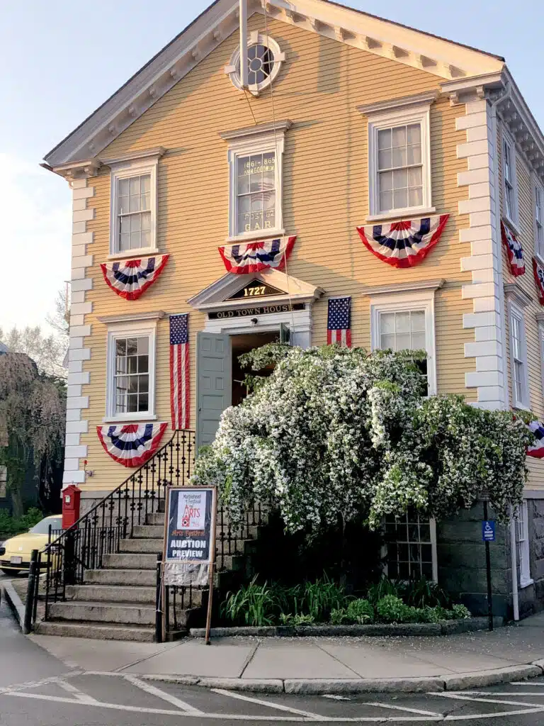A yellow historic building with patriotic bunting and American flags, flowering tree, and a sign by the steps.