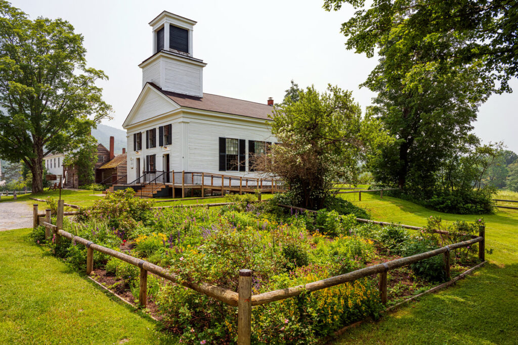 A white wooden church with a bell tower stands behind a small fenced garden on a sunny day.