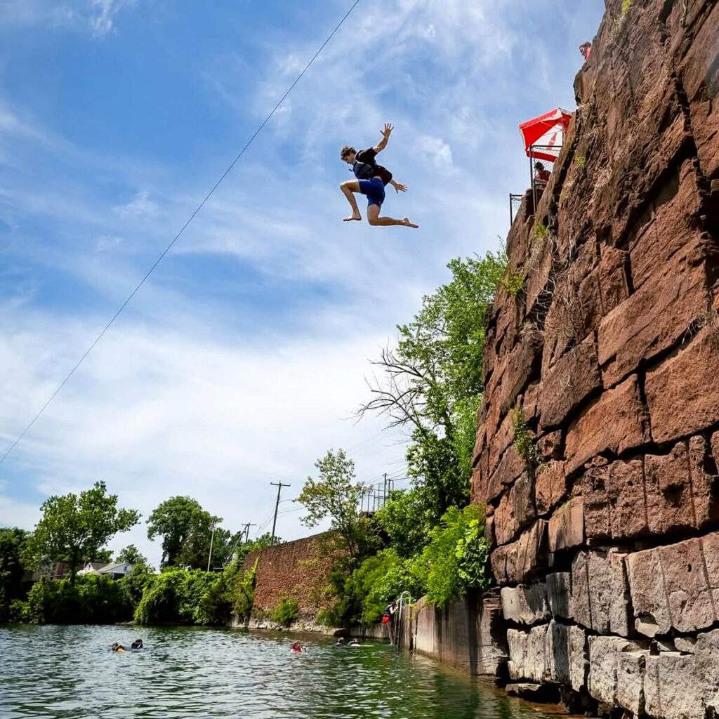 Best New England Summer Attractions for Families. Person mid-air jumping off a rocky cliff into a river, with trees and bright sky in the background.