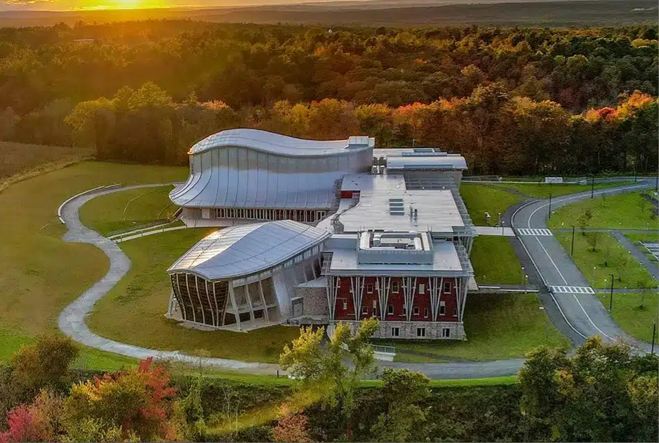 Groton Hill Music Center, an architectural and acoustical wonder in Groton, MA. Modern building with curved roofs surrounded by trees and grass at sunset, viewed from above.