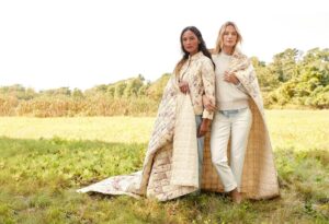 Two women standing in a grassy field, one in a quilted coat, the other wrapped in a quilt, both looking at camera.