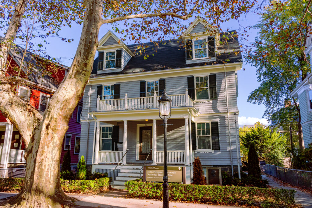 A three-story gray house with white trim, a front porch, and trees lining the sidewalk in front.