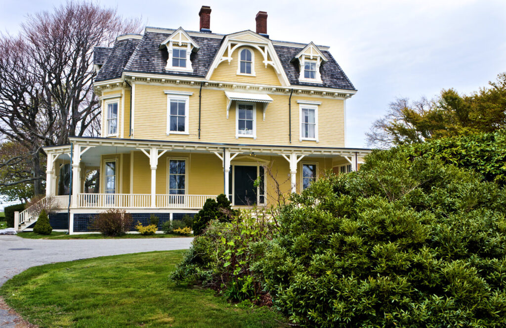 Large yellow Victorian house with a mansard roof, white trim, and wraparound porch, surrounded by green bushes.