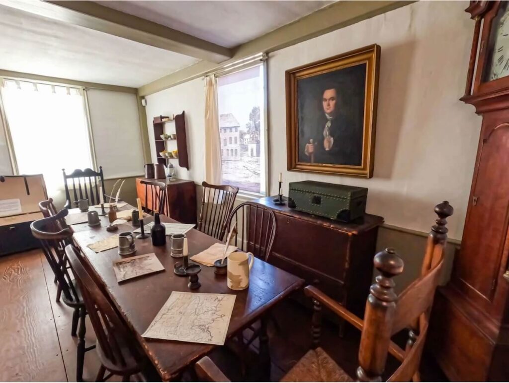 A historic room with a wooden table, chairs, maps, mugs, and a portrait hanging on the wall.