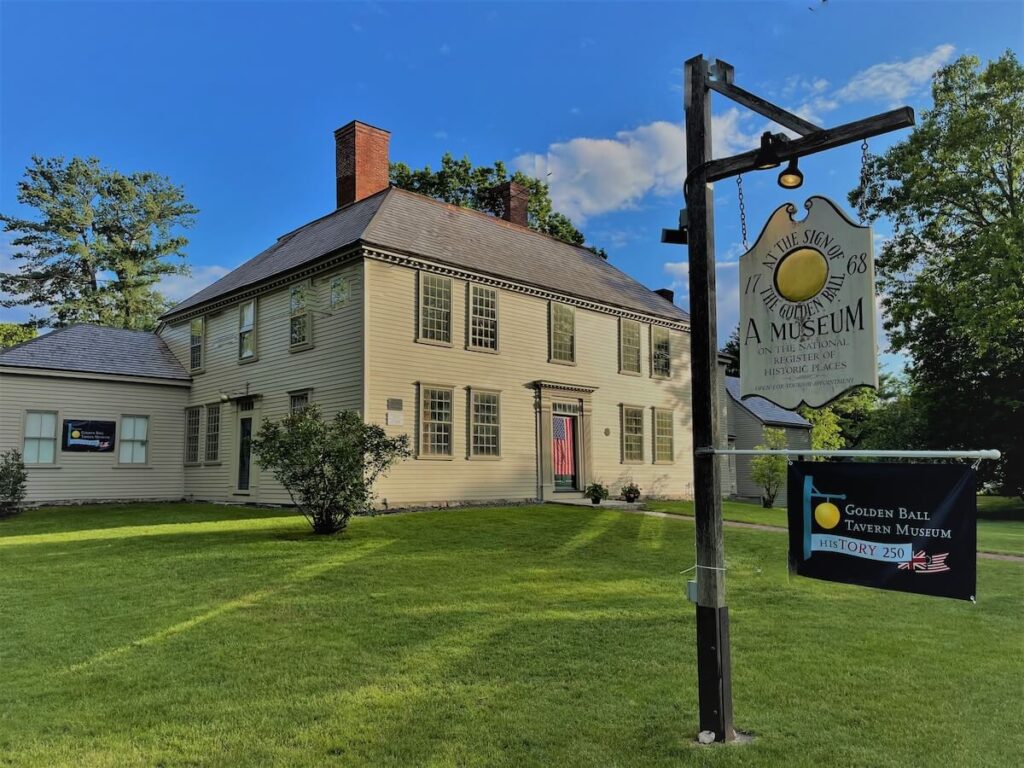 A historic two-story house with a sign reading "Golden Ball Tavern Museum" stands on a sunny, green lawn.